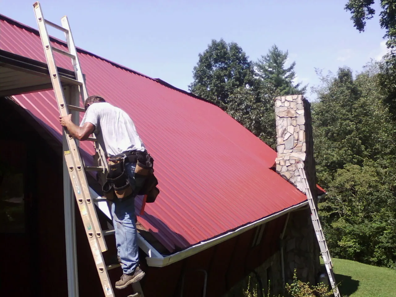 Roofer on a ladder beside a red metal roof and stone chimney — Soffit & Fascia Repair by Roofing Brothers in Bullock